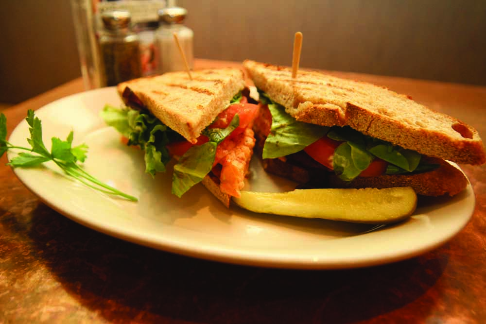 A grilled salmon B.L.T. sandwich on a dinner plate with a pickle and a garnish from Cafe Bernardo in downtown Sacramento. (Photo by Bryce Fraser)