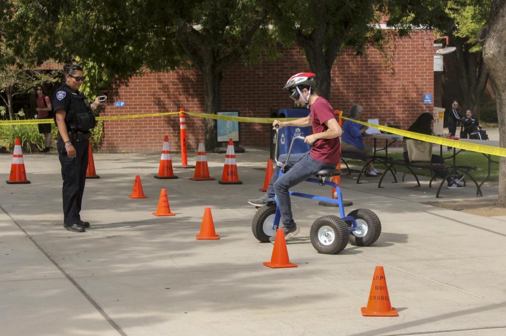 Allen Marinovich, Chemistry major, tries to make a turn with a tricycle while wearing alcohol impairment simulation goggles. Officer Rosie Salazar from Los Rios Police department makes sure students are safe during the simulation. (Photo by Itzin Alpizar) 