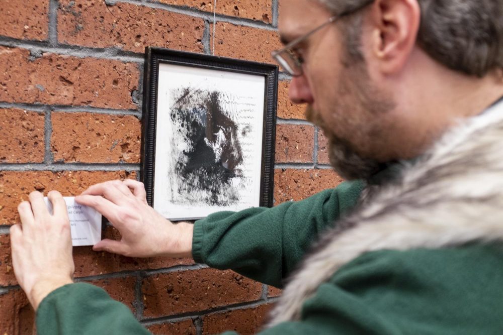 Brad Carps, studio art major, hangs a nameplate besides a monoprint of his which hangs in the American River College library stairwell. The piece titled “Epilepsy” is one in a semester-long show of student work by members of the Art Club at ARC, a collective of sudents on campus with a common interest of creating artwork. Carps is the president of the art club and organized the show which includes work by Max Marchol, Frankie Vanity, Clare and Carolan Korten and other members of the Art Club at ARC. (Photo by Patrick Hyun Wilson)