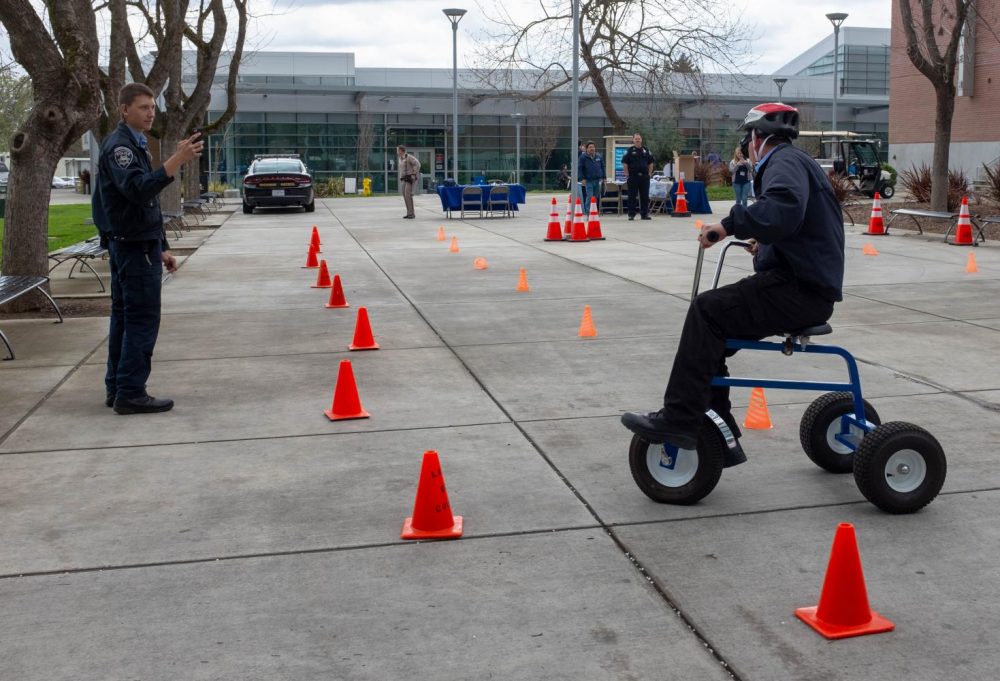 Outside the library at American River College, campus patrol officer John Lawyer rides a tricycle while wearing drunk goggles for the California Highway Patrol DUI Awareness tricycle obstacle course on March 20, 2019. (Photo by Patrick Hyun Wilson)
