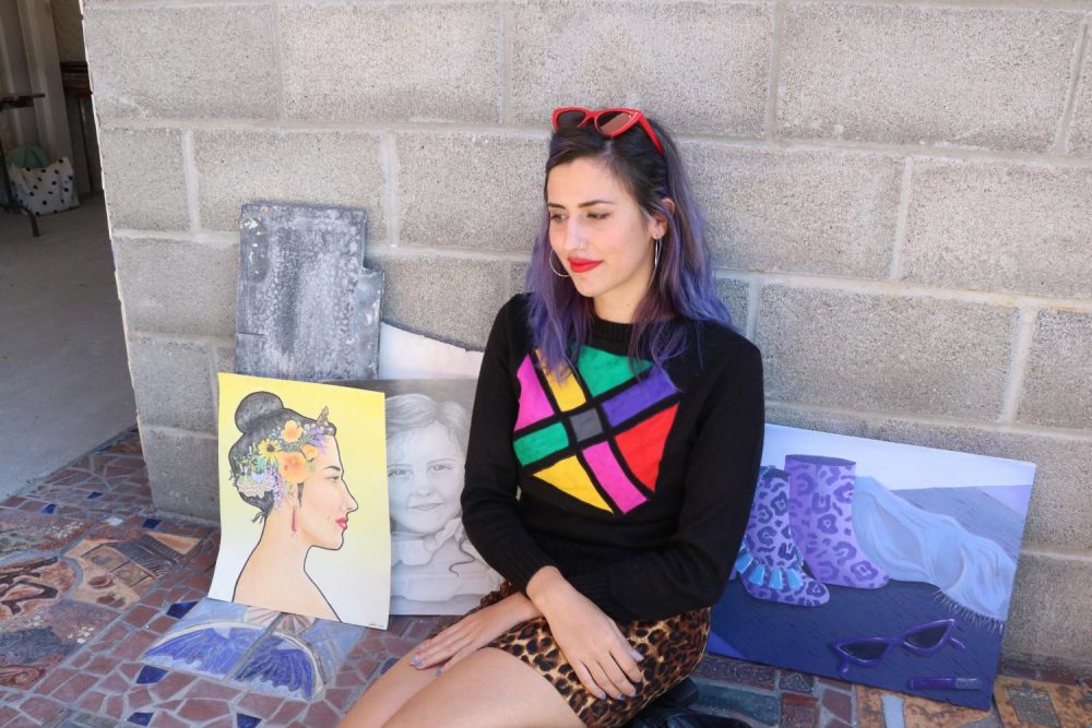 Art major Cat Alfstad sits between the art buildings among her own art (self portrait titled Cat (left), Lucy Rose (middle) and But Make it Fashion (right) at American River College on Oct. 17, 2019. (Photo by Ariel Caspar)